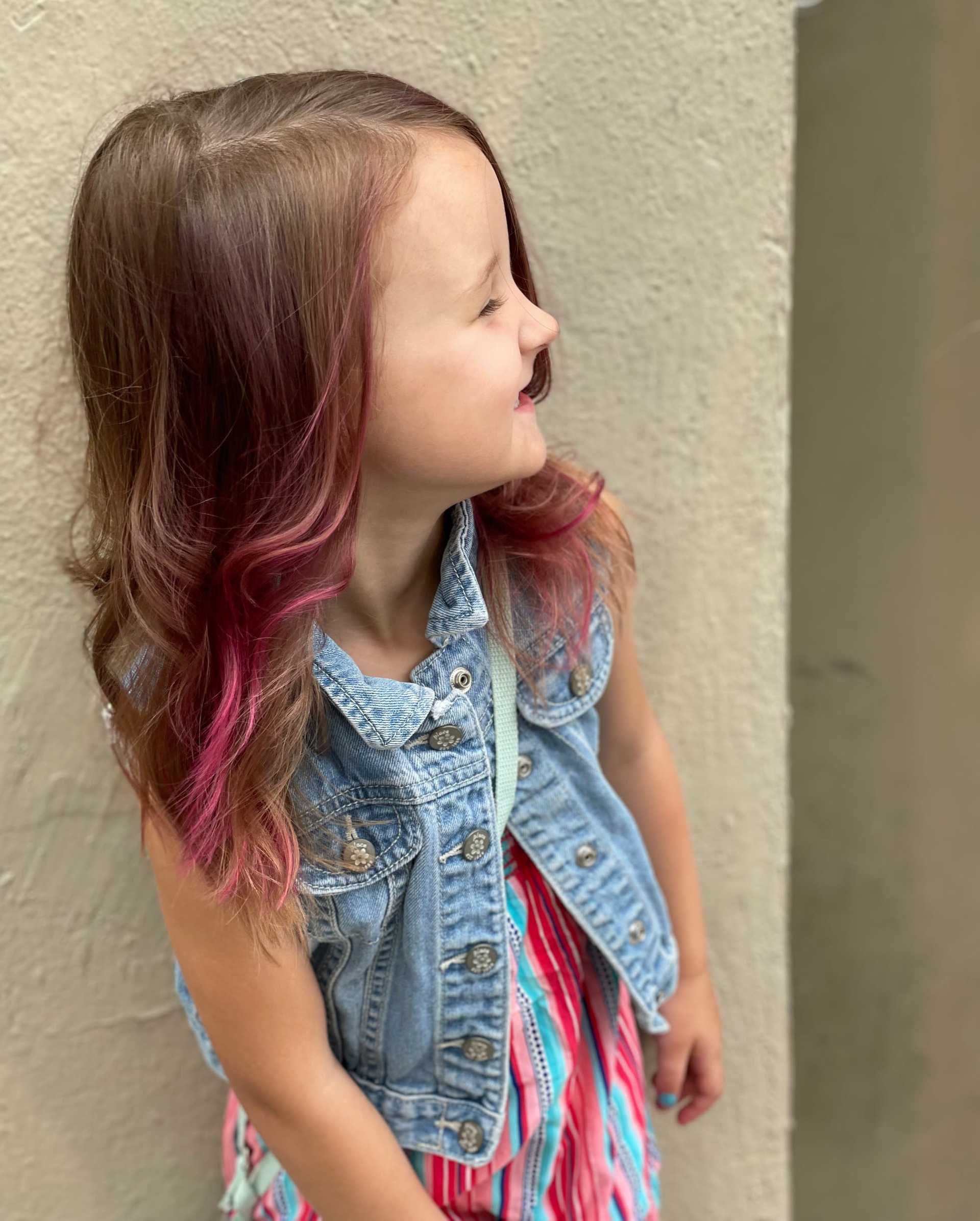 Young girl with pink streaks, denim vest, and colorful dress smiles.