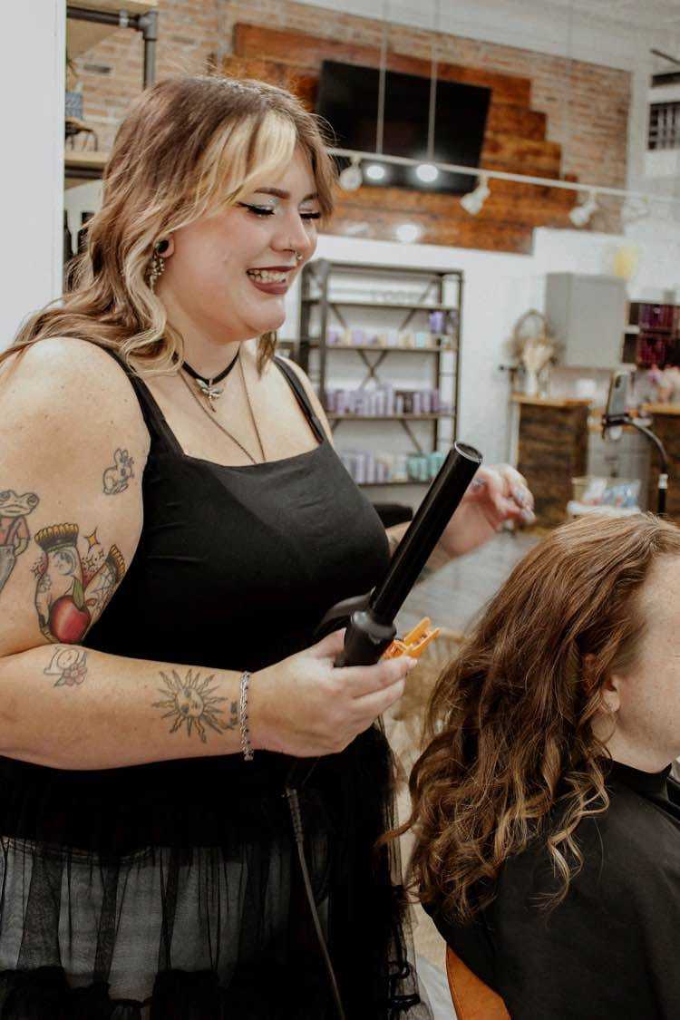 Hairdresser with tattoos styles a client's curly hair using a curling iron in a salon.