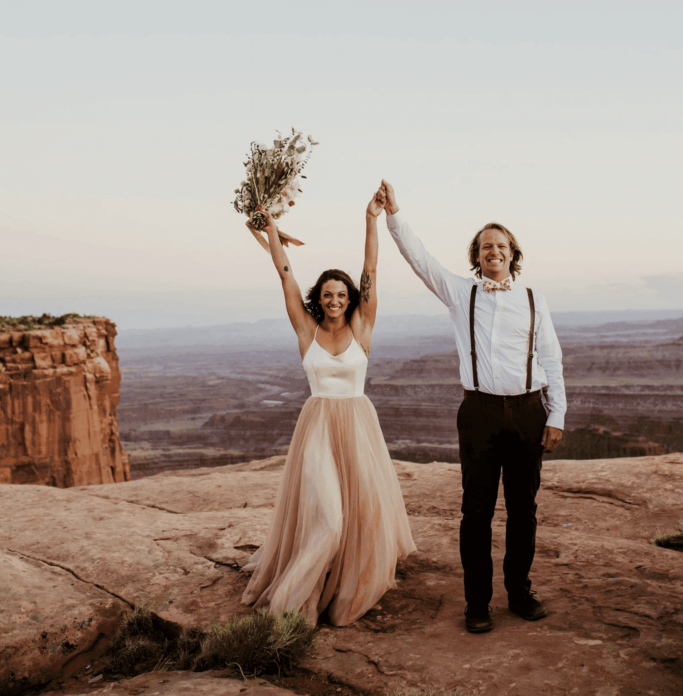 Bride and groom celebrating on a rocky cliff with a scenic canyon view in the background.